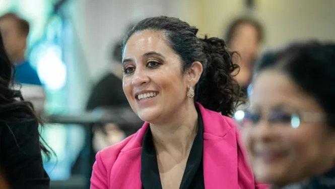 membership A woman in a gray blazer is attentively listening and engaging in a conversation at a professional event.