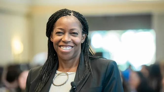 sponsorship A woman with braided hair, wearing a black blazer, smiles confidently at the camera in a well-lit conference room.